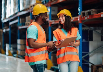 Two smiling warehouse workers wearing safety vests and hard hats are discussing logistics